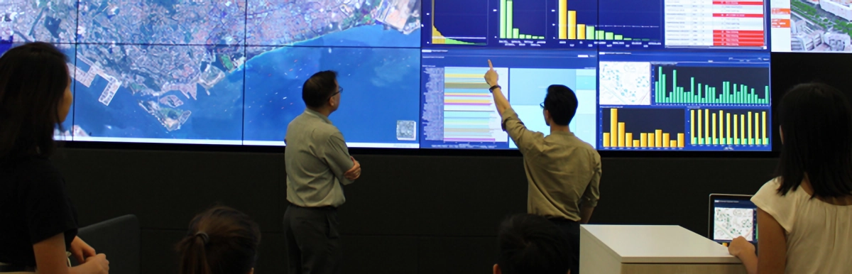 People stand before a large display wall showing satellite maps and colorful data charts, as one person points at the screen.